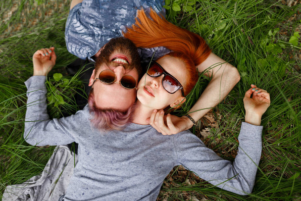 Радісна пара, сидячи на зеленій траві, в парку, сидячи з повернутими головами, в сонцезахисних окулярах, дивлячись на камеру. Зміст стилю життя
. - Фото, зображення