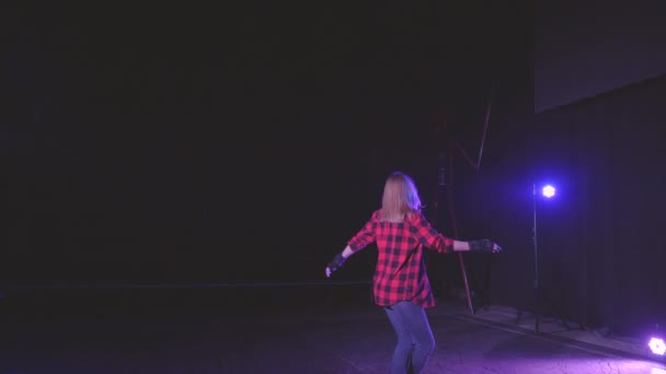 Blonde roller girl with protective gloves, gracefully swirls on rollerblades on front wheels in dark studio under neon pink blue light of spotlights in slow motion. concept active sports lifestyle - Footage, Video