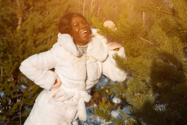 Femme à la peau foncée vêtue d'un manteau de fourrure blanche dans une forêt hivernale ensoleillée - Photo, image