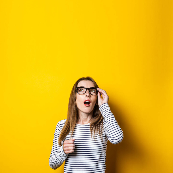 Young woman looks up in surprise, holding on to glasses on a yellow background - Photo, Image
