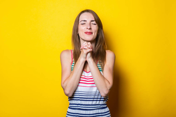 Young smiling woman with closed eyes folded hands together, on yellow background - Photo, image