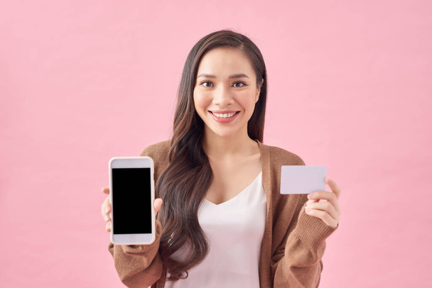 Πορτρέτο μιας χαμογελαστής νεαρής γυναίκας που κρατά λευκό οθόνη κινητό τηλέφωνο, που δείχνει την πιστωτική κάρτα απομονώνονται σε ροζ φόντο - Φωτογραφία, εικόνα