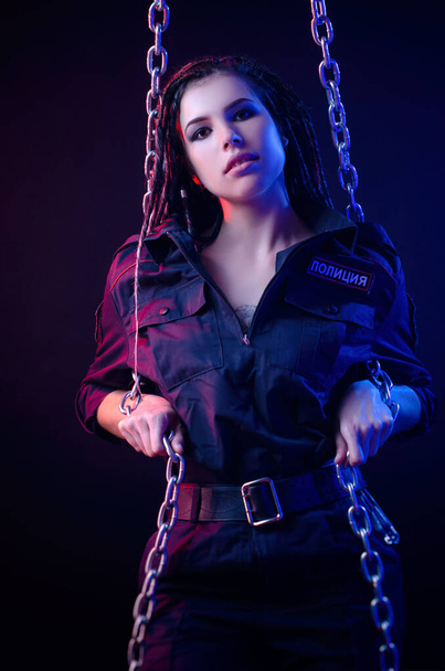 a girl in a police uniform with dreadlocks in neon light with chains and handcuffs English translation the police - Foto, Bild