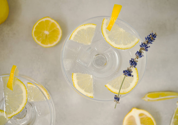 Clear water in a glass with ice and lemon slices stands on a light gray concrete background. The glass is decorated with a sprig of lavender and a slice of lemon. Near are sliced lemons. Top view, close up - Valokuva, kuva