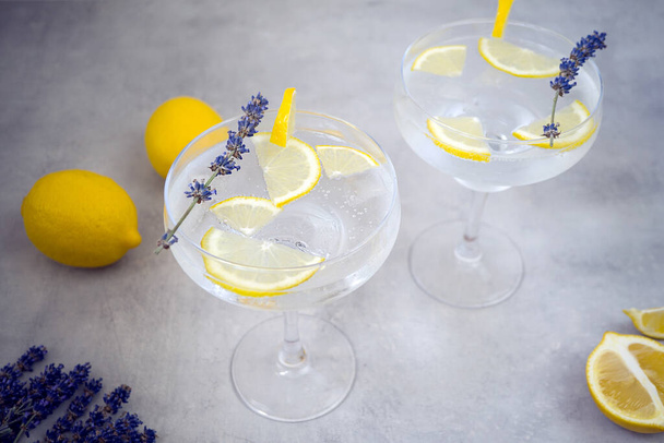 Lemon non-alcoholic cocktails with ice cubes in beautiful glasses for margaret stands on a gray concrete background. Glasses are decorated with lavender and a slice of lemon. Nearby lies two lemons. - Foto, Imagen