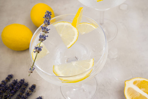 Infused water with ice and lemon in a glass stands on a gray concrete background. The glass is decorated with a sprig of lavender and a slice of lemon. Nearby are two lemons. Close-up, top view - Foto, Imagen