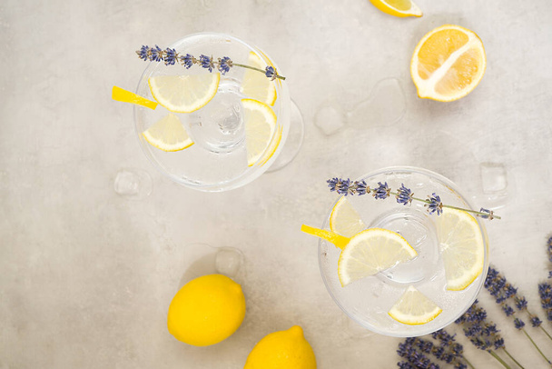 Two glasses of citrus infused water with ice and lemon slices stand on a gray concrete background. Near are two lemons, lavender flowers and melting ice cubes. Top view, vertically - Φωτογραφία, εικόνα
