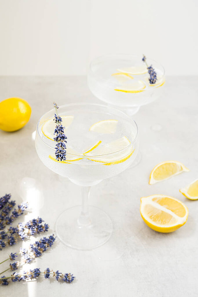 Two glasses of summer cool drinks with fresh lemon stand on a gray stone table. Glasses are decorated with purple lavender flowers. Nearby lie slices of lemon and lavender. Vertically - Zdjęcie, obraz