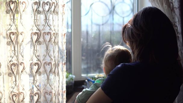 Een jonge moeder houdt een baby in haar armen. Een vrouw met een kind dat uit het raam kijkt en het gordijn dicht duwt.. - Foto, afbeelding