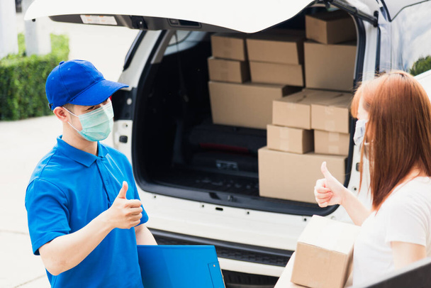Asian delivery express courier young man giving parcel boxes to woman customer receiving both protective face mask and show thumbs up finger for good support sign, under curfew pandemic coronavirus - Foto, Imagem