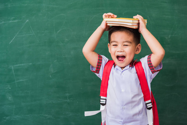Back to School. Happy Asian funny cute little child boy from kindergarten in student uniform with school bag and book on head smiling on green school blackboard, First time to school education concept - Photo, Image