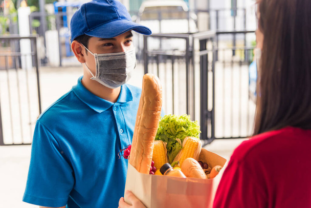 Asian young delivery man in uniform wear protective face mask he making grocery service giving fresh food to woman customer receiving front house under pandemic coronavirus, Back to new normal concept - Фото, изображение