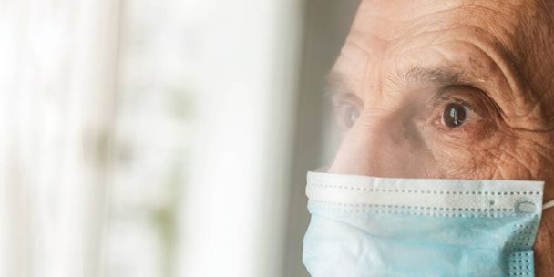 sad pensioner in blue sterile mask looks through window - Fotó, kép