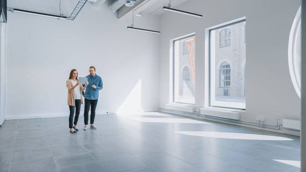 若いヒップスター男性と女性は空のホワイトオフィスに立って、タブレット上の拡張現実ソフトウェアでそれをマップします。大きな窓から差し込む陽射し. - 写真・画像