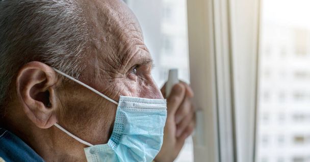 old man in blue disposable face mask looks through window - Valokuva, kuva