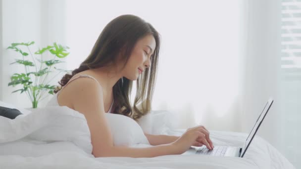 Beautiful Attractive Asian young woman lying on bed and using computer laptop for using social media and shopping online relax in cozy bedroom in holiday at home,Lifestyle in bedroom Concept - Felvétel, videó