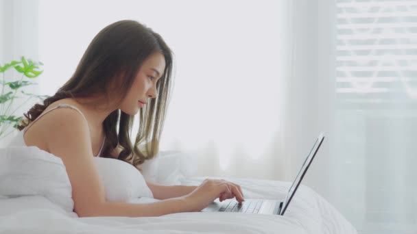 Beautiful Attractive Asian young woman lying on bed and using computer laptop for using social media and shopping online relax in cozy bedroom in holiday at home,Lifestyle in bedroom Concept - Materiał filmowy, wideo
