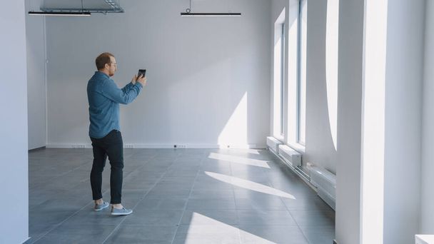 Young Hipster Man in Jeans Shirt Standing in Empty Office and Map it with an Augmented Reality Software on a Tablet. Sunlight Shines Through Big Windows. - Fotoğraf, Görsel