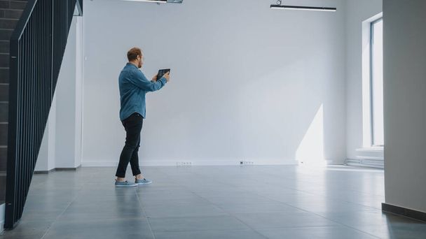 Young Hipster Man in Jeans Shirt Standing in Empty Office and Map it with an Augmented Reality Software on a Tablet. Sunlight Shines Through Big Windows. - Foto, Imagen
