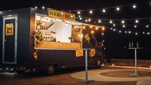 Empty Scene with a Dark Street Food Van Standing in the Evening in a Nice Warmly Lit Neighbourhood Next to the Sea. Food Truck Has Burgers and Drinks for Sale. Tables Have Bottles on Them. - 写真・画像