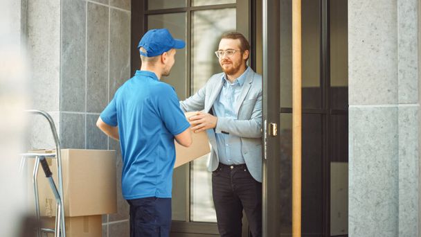 Delivery Man Gives Postal Package to a Business Customer. In Stylish Modern Urban Office Area Courier Delivers Cardboard Box Parcel to a Man - Фото, зображення