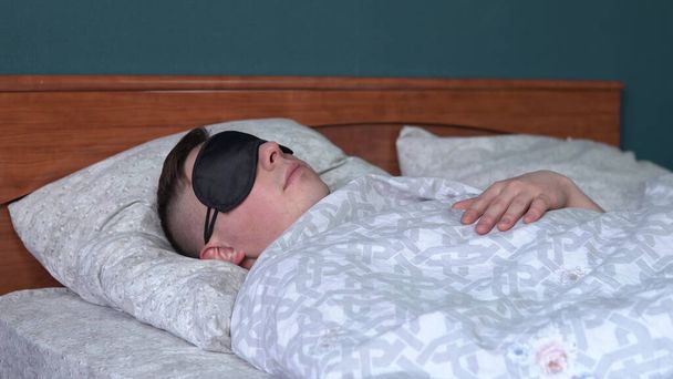 Un joven duerme con una máscara para dormir. Un hombre yace en la cama en su habitación
. - Foto, Imagen