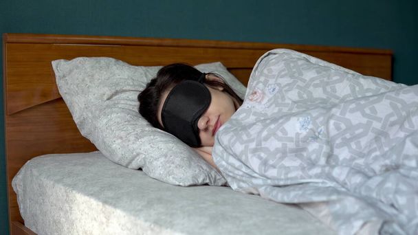Μια νεαρή γυναίκα κοιμάται με μάσκα ύπνου. Ένα κορίτσι ξαπλώνει στο κρεβάτι στο δωμάτιό του.. - Φωτογραφία, εικόνα