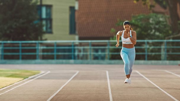 Schöne Fitness-Frau in hellblauem Sporttop und Leggings startet einen Sprintlauf in einem Freiluftstadion. Sie läuft an einem warmen Sommertag. Athletin macht ihre Sportpraxis. - Foto, Bild