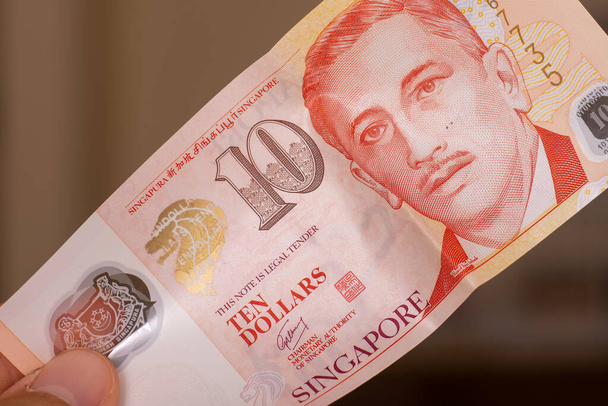 Osoby strony podając walutę Singapuru - Jeden czerwony dziesięć dolarów banknotu rozłożone na brązowym tle. Wymiana pieniędzy. - Zdjęcie, obraz