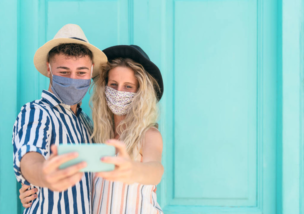 Young couple wearing face mask taking selfie with mobile smartphone on vacation - People having fun traveling again during corona virus outbreak - Love relationship and technology concept - Photo, Image
