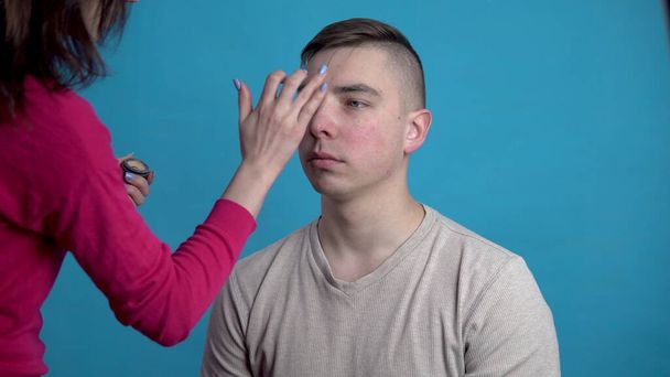 El maquillaje se aplica a un joven. La chica aplica maquillaje a un hombre antes de disparar sobre un fondo azul
. - Foto, Imagen