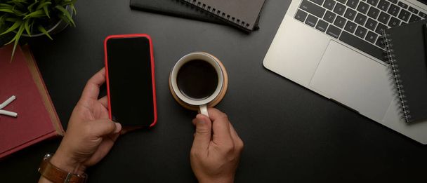 Nad hlavou záběr mužského podnikatele levé ruky pomocí smartphonu, zatímco pravá ruka drží šálek kávy na tmavém moderním kancelářském stole - Fotografie, Obrázek