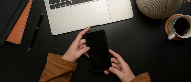 Overhead tiro de mãos segurando smartphone no espaço de trabalho moderno escuro com laptop, suprimentos e decoração
 - Foto, Imagem