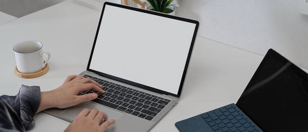 Plan recadré des mains féminines à l'aide d'un ordinateur portable simulé sur un bureau à domicile blanc avec tablette numérique et tasse à café  - Photo, image
