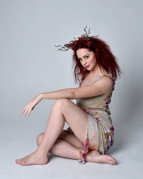 full length portrait of red hair girl wearing floral dress and flower head dress. sitting pose on a grey studio background. - Foto, imagen