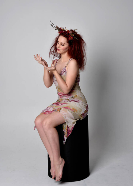 pose pleine longueur de fille aux cheveux rouges portant une robe de fée florale et une coiffure. pose assise sur fond de studio gris. - Photo, image