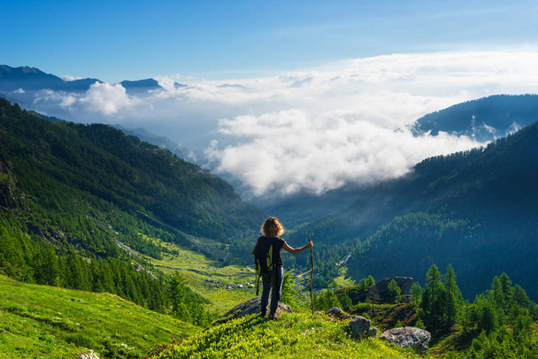 Woman resting on mountain top, looking at view dramatic landscape clouds over the valley, clear blue sky. Summer activity fitness wellbeing freedom success. - Valokuva, kuva