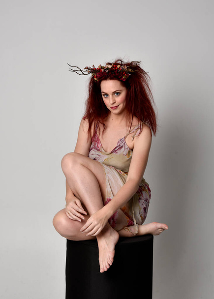 full length pose of red hair girl wearing floral fairy dress and headdress. sitting pose on grey studio background. - Фото, зображення