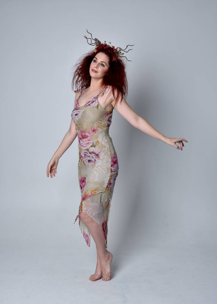 full length portrait of red haired girl wearing floral dress and headdress. standing pose on grey studio background. - Фото, изображение