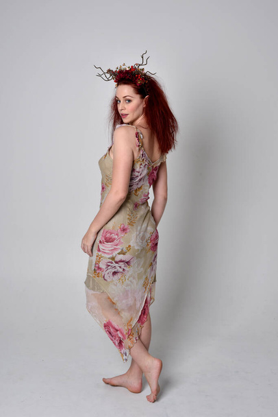 full length portrait of red haired girl wearing floral dress and headdress. standing pose on grey studio background. - Photo, Image