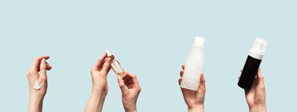 Hands up holding beauty cosmetic products isolated on blue background horizontal banner format. Woman takes oil serum, toner or tonic bottle, facial foam cleanser and smear smudge moisturizer cream - 写真・画像