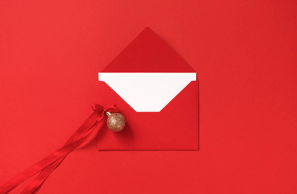 Kırmızı zarf ve kırmızı arka planda Atlas kurdelesinin yayıyla altın parıltılı Noel topu. Minimalist yeni yılı kutluyoruz. Parlak ve zarif bir model. - Fotoğraf, Görsel