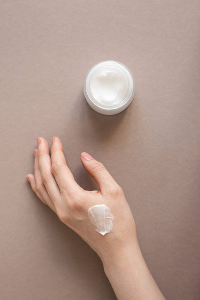 Cream moisturizer smudge apply on woman hand with nude pink manicure on nails and body lotion in glass jar on beige background top view, vertical. Beauty skincare concept - Photo, Image