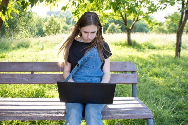 A young woman or teenage girl with a leptop on a bench is  surfing the Internet or studying outdoors. Holidays, summer mood, Sunny summer day. - Φωτογραφία, εικόνα