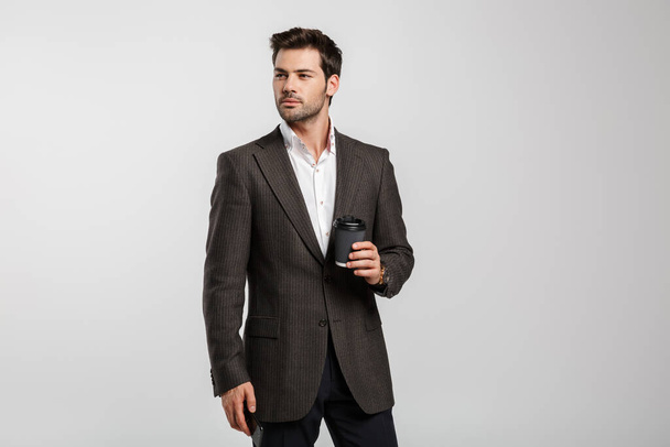 Image of young successful businessman in jacket drinking coffee takeaway and holding cellphone isolated over white background - Foto, immagini