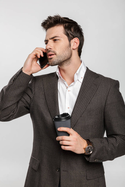 Image of young serious businessman in jacket drinking coffee takeaway and talking on cellphone isolated over white background - Valokuva, kuva