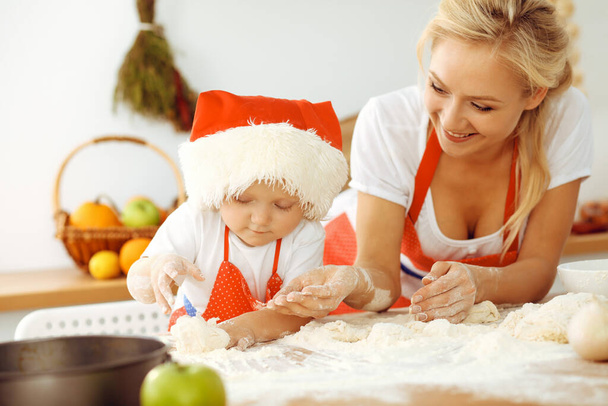 Little girl and her blonde mom in red aprons playing and laughing while kneading the dough in kitchen. Homemade pastry for bread, pizza or bake cookies. Family fun and cooking concept - Фото, изображение