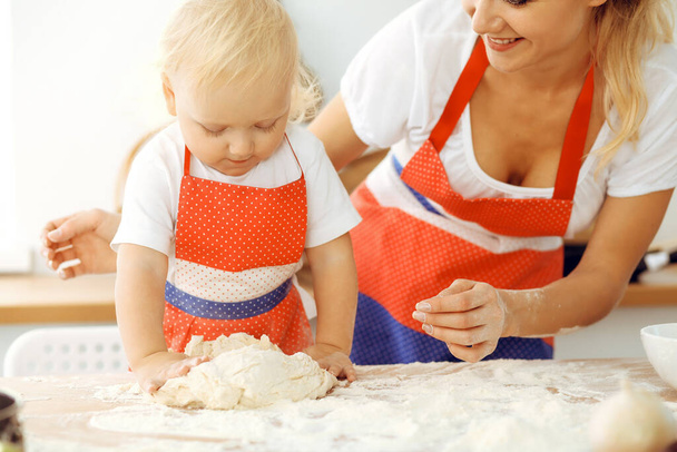 Little girl and her blonde mom in red aprons playing and laughing while kneading the dough in kitchen. Homemade pastry for bread, pizza or bake cookies. Family fun and cooking concept - Zdjęcie, obraz