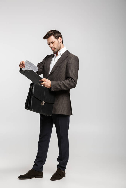 Image of a concentrated serious young businessman isolated over white wall background holding bag and clipboard. - Foto, afbeelding