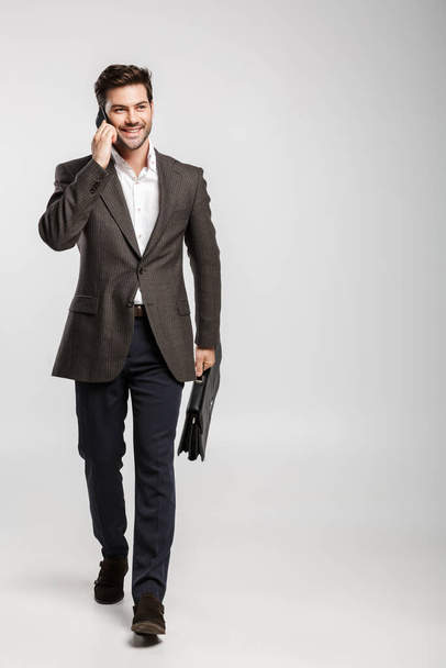 Image of young smiling businessman in jacket talking on cellphone while walking with bag isolated over white background - Foto, Bild